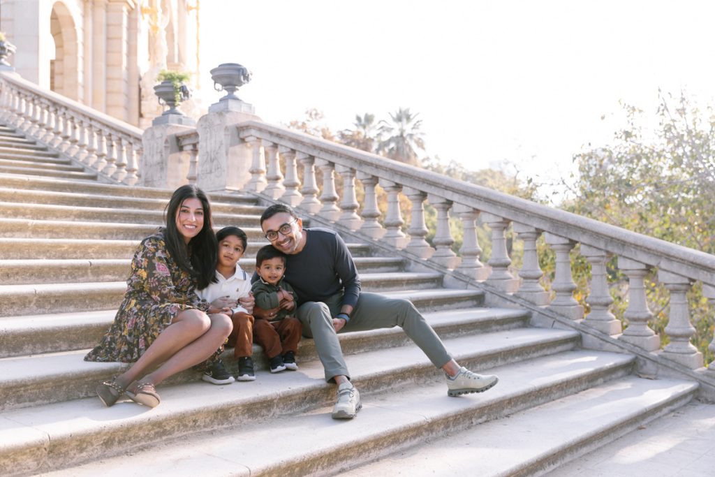A smiling family of four, including two young boys, sits together on stone steps outdoors, with a historic building and trees in the background.