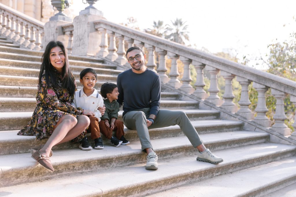 A smiling family of four sits together on stone steps outdoors. Two parents and two young boys, dressed casually, pose in front of a balustrade with trees and a building visible in the background.