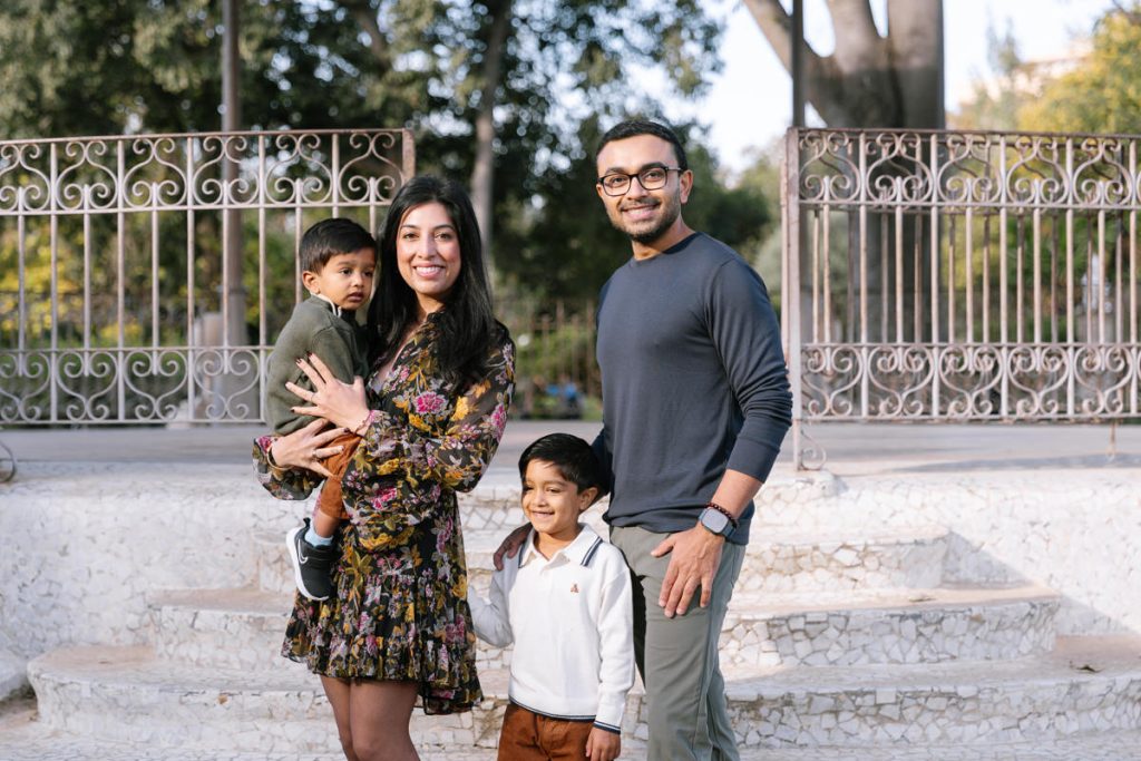 A family of four poses outdoors, smiling at the camera. The mother holds a young child, while the father stands beside her with an older child in front of him. Trees and a decorative metal fence are in the background.