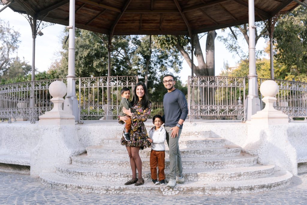 A family of four poses and smiles on stone steps in front of a round gazebo in a park. The mother holds a young child, while the father stands next to an older child. Trees and iron railings are visible in the background.