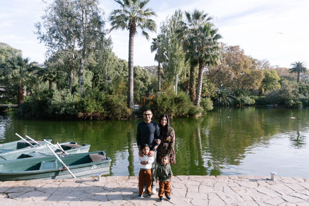 A family of four stands by a lakeside with two small boats docked nearby. Behind them are lush green trees and palm trees reflected in the water under a clear sky.