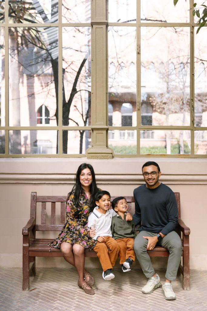 A smiling family of four sits on a wooden bench indoors by a large window. The mother and father flank their two young sons, who sit in the middle laughing. Sunlight streams through the window behind them.