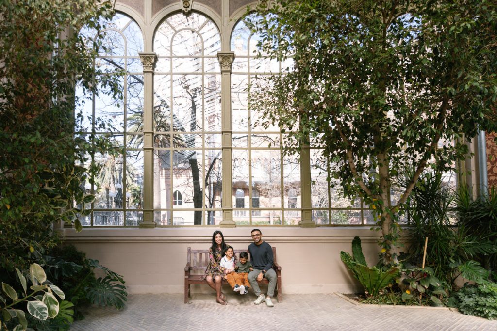 A family of four sits on a bench inside a bright conservatory with large arched windows and lush green plants all around them. The view outside shows trees and a historic building.