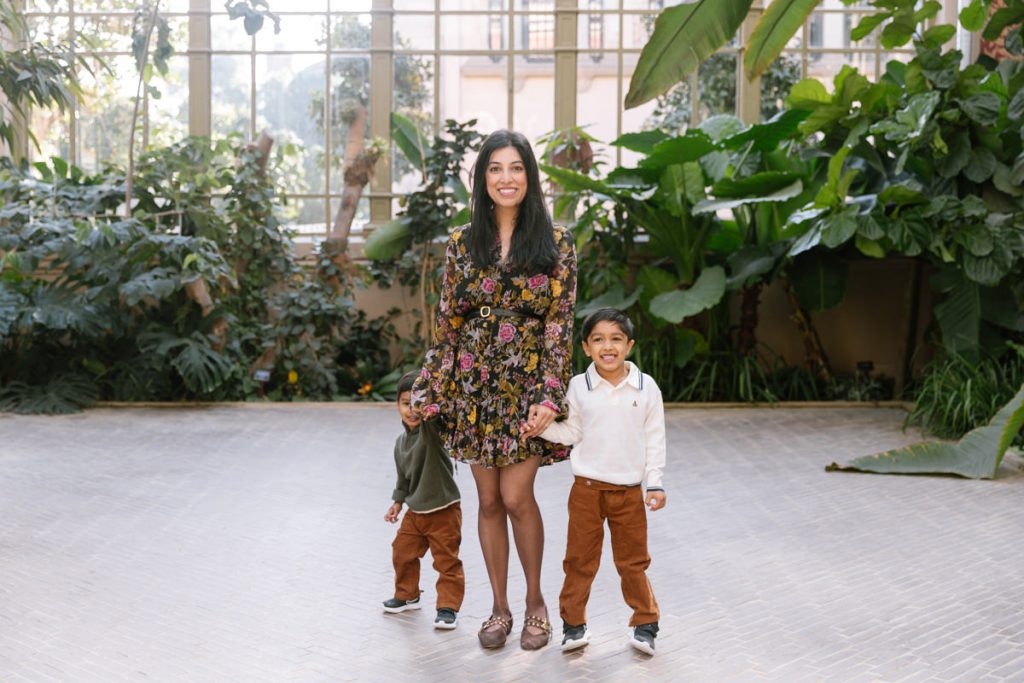 A woman in a floral dress stands smiling indoors, holding hands with two young boys. Both boys wear brown pants; one wears a green shirt and the other a white sweater. They are surrounded by lush green plants.