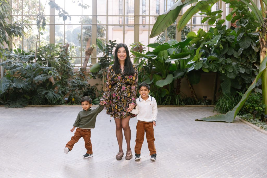 A woman in a floral dress stands indoors holding hands with two young boys in brown pants. They are smiling and surrounded by lush green plants with large windows in the background.