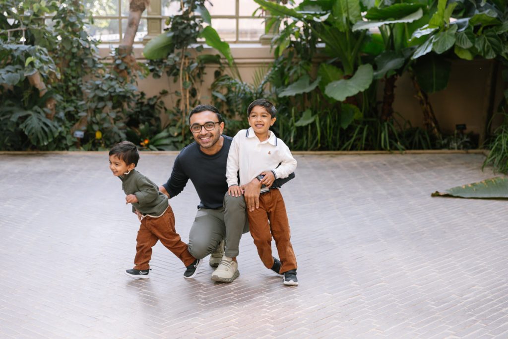 A man wearing glasses squats and smiles, hugging two young boys. All three are smiling. The boys wear matching brown pants, and they are surrounded by lush green plants indoors.
