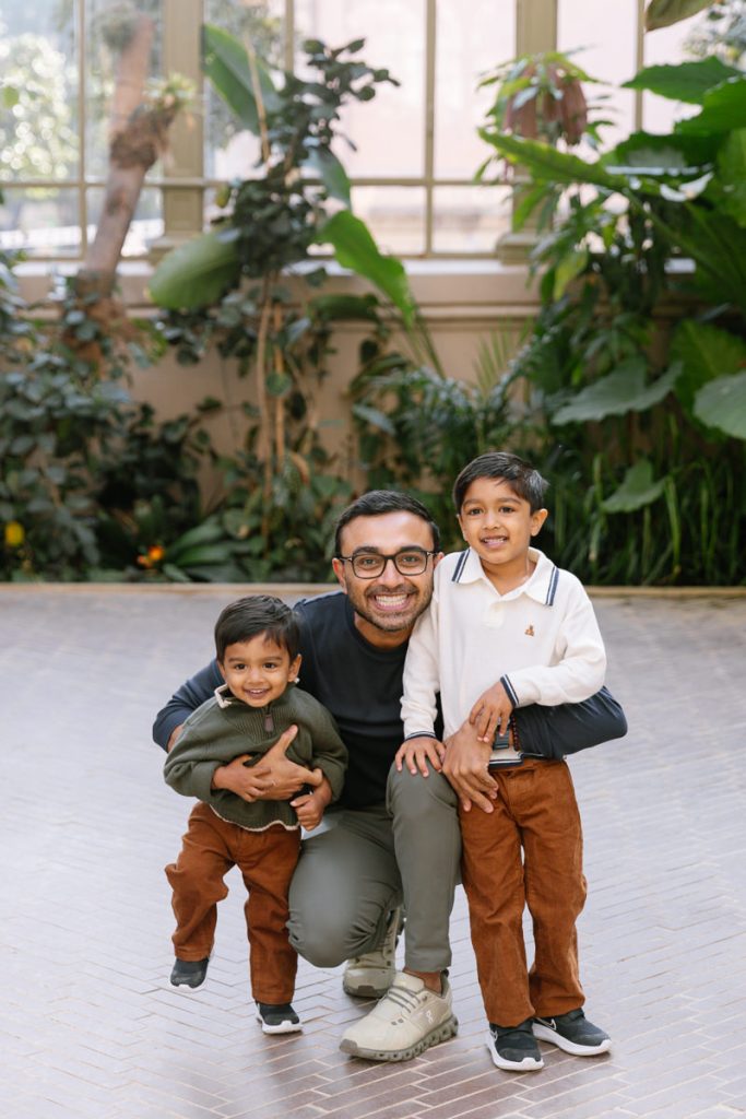 A smiling man kneels indoors with his arms around two young boys. All three wear glasses and casual clothes. The boys wear brown pants and sweaters, and large green plants fill the background.