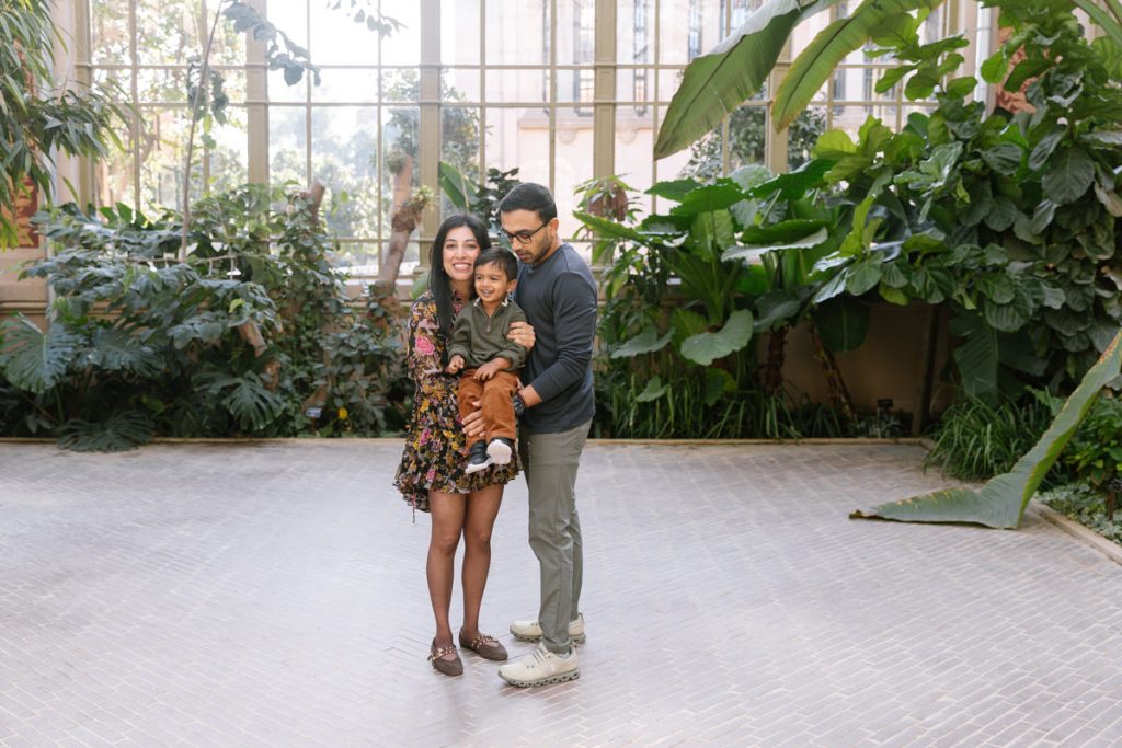A smiling family with a woman, man, and young child stands together indoors, surrounded by large green plants and tall windows that let in natural light.