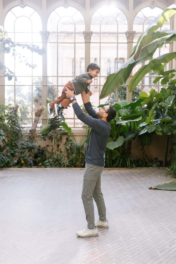 A man lifts a smiling child into the air inside a bright, spacious room with large windows and lush green plants. The scene conveys joy and playfulness.