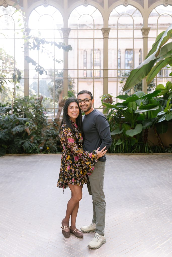 A smiling couple stands close together in a bright, indoor garden with tall arched windows, lush green plants, and sunlight streaming in behind them. The woman wears a floral dress and the man wears a dark shirt and light pants.