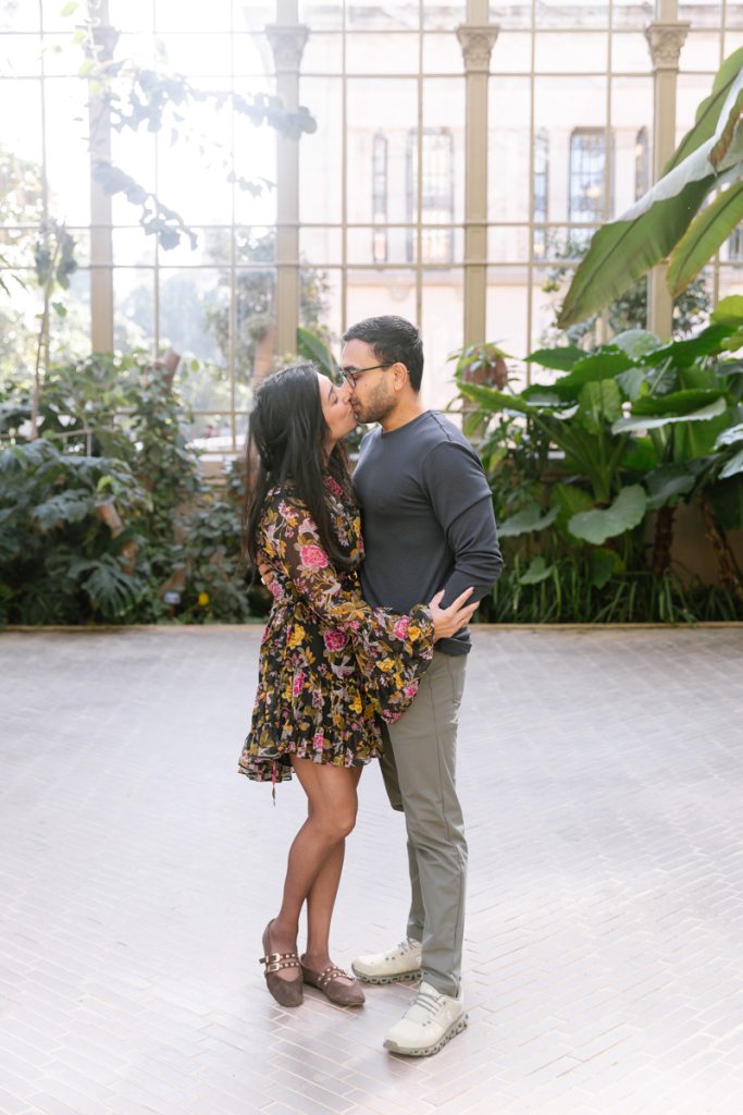 A couple stands closely together, embracing and sharing a gentle kiss in a bright conservatory filled with lush green plants and natural light streaming through large windows.