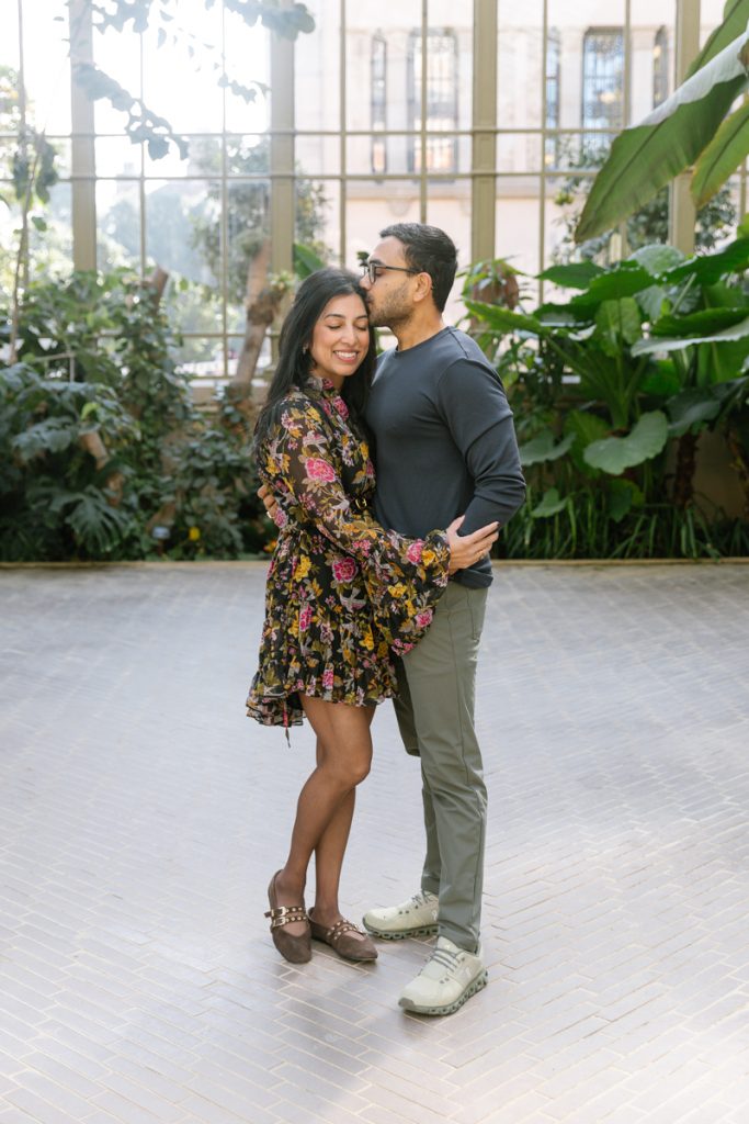 A man kisses a woman on the forehead as they stand close together in a bright, indoor garden. The woman wears a floral dress and smiles, while the man wears glasses and casual clothes. Lush greenery surrounds them.