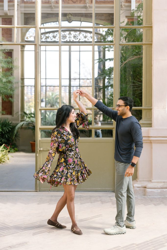 A man twirls a smiling woman in a floral dress inside a bright, elegant room with tall glass doors and potted plants, creating a joyful, playful moment together.