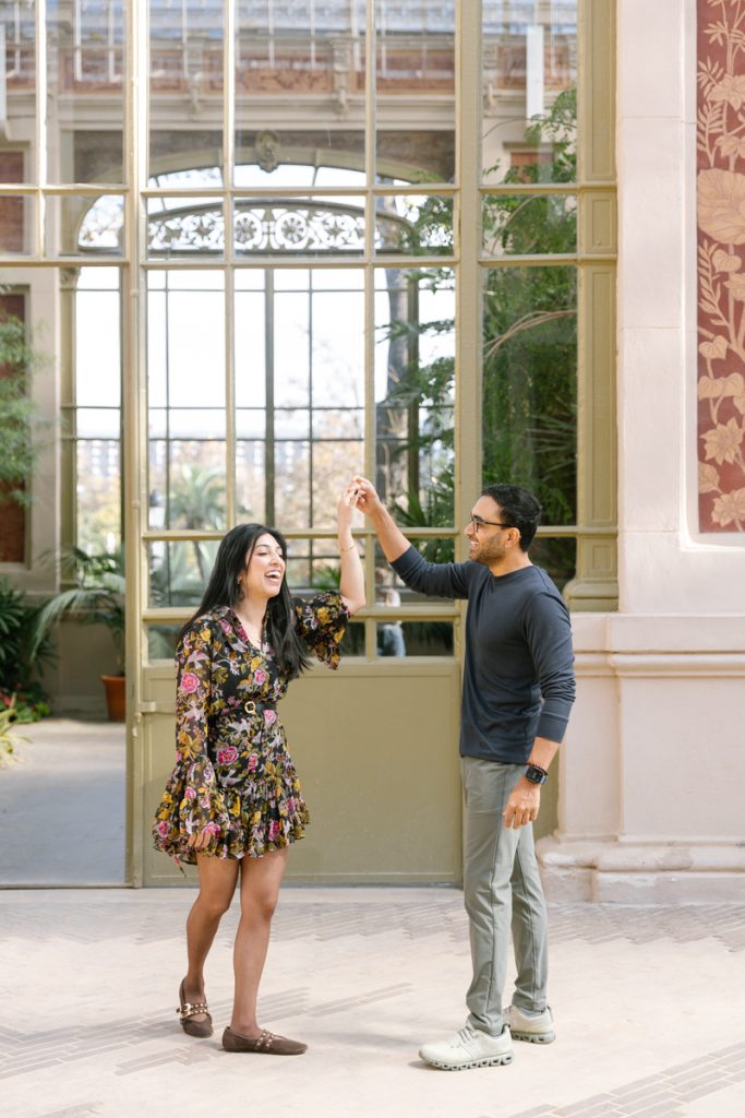 A smiling couple dances together indoors; the woman wears a floral dress and the man wears a dark shirt and light pants. They hold hands, standing in front of large windows with plants visible in the background.