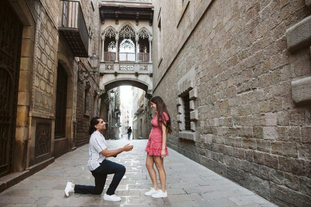 A man kneeling on his knee to a woman in a red dress during a surprise engagement in front of Pont del Bisbe in the Gothic Quarter in Barcelona.