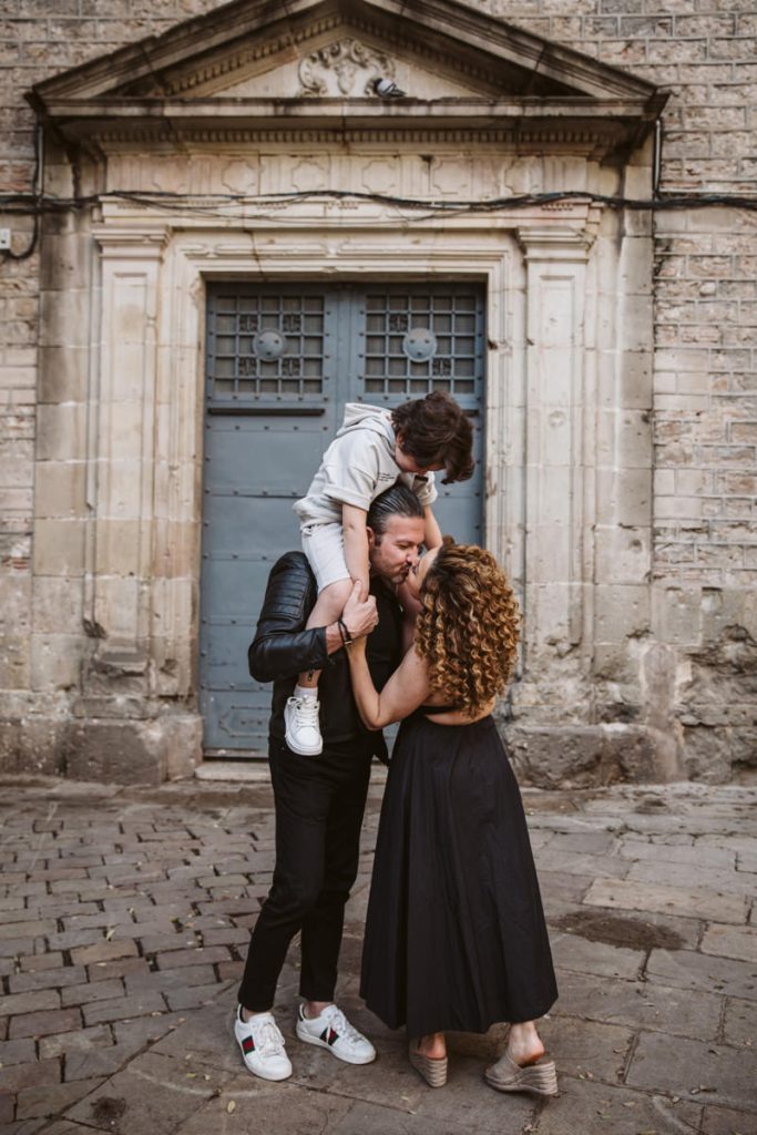 A man holds a child on his shoulders while a woman in a black dress kisses the child’s cheek, capturing sweet family photos in the Gothic Quarter in Barcelona, in front of a historic stone building with a blue-gray door.