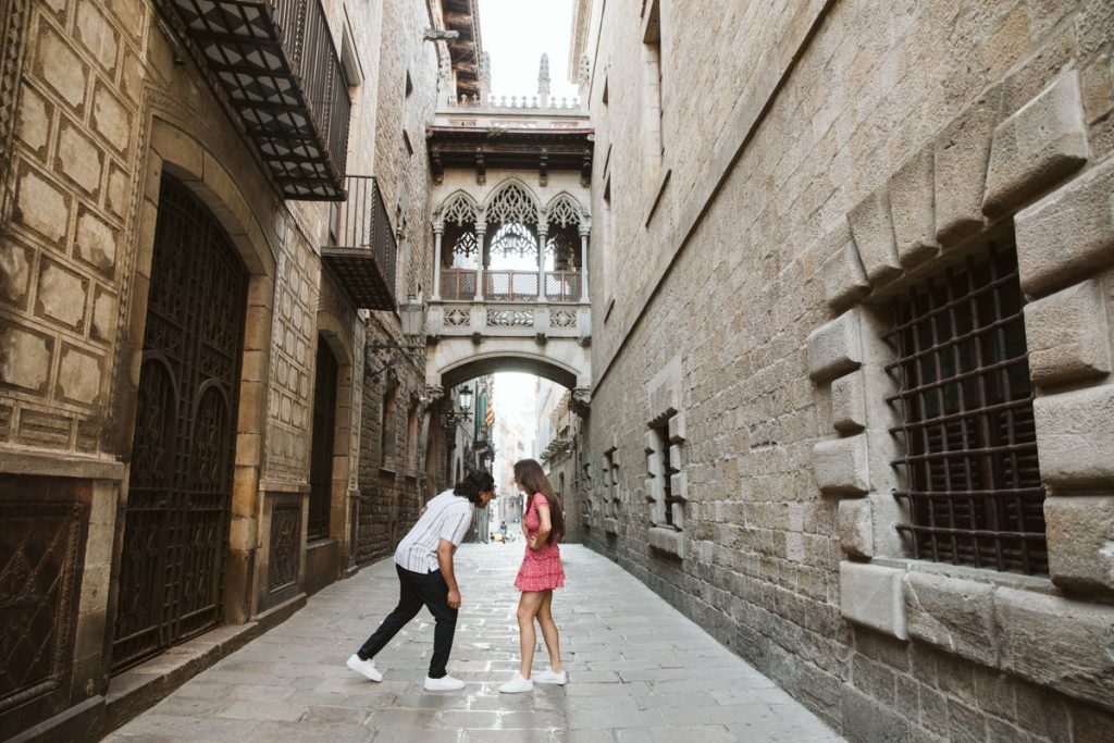 Two people playfully interact on a narrow cobblestone street during a surprise proposal in front of Pont del Bisbe in the Gothic Quarter in Barcelona, surrounded by historic stone buildings with intricate windows and an arched bridge above.