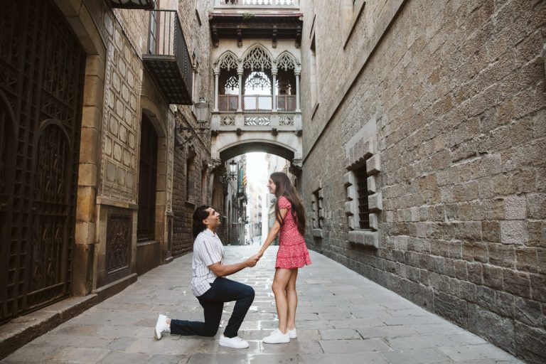 A man kneels and holds hands with a surprised woman in a cobblestone alley, set among historic stone buildings and an ornate bridge—a romantic and joyful surprise proposal in the Gothic Quarter in Barcelona.