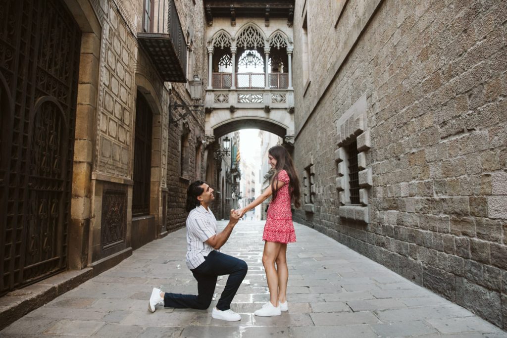 A man kneels for a surprise proposal in the Gothic Quarter in Barcelona, asking a woman in a pink dress to marry him on a cobblestone street amid historic stone buildings and an ornate arched bridge.