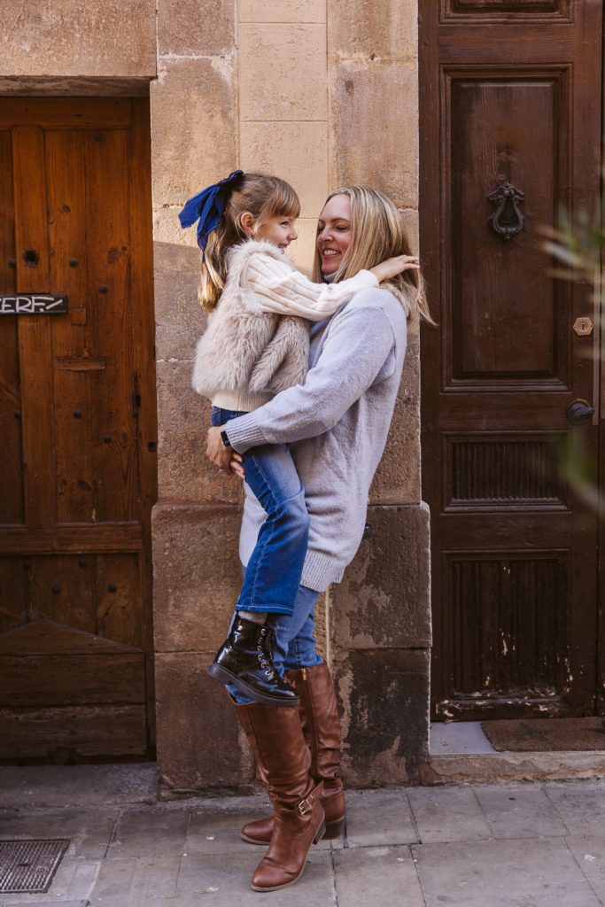 A woman in a light sweater and brown boots smiles while holding a young girl with a blue bow, furry vest, jeans, and boots. They are on a stone street by wooden doors during a family photo session in Vila de Gràcia, Barcelona.