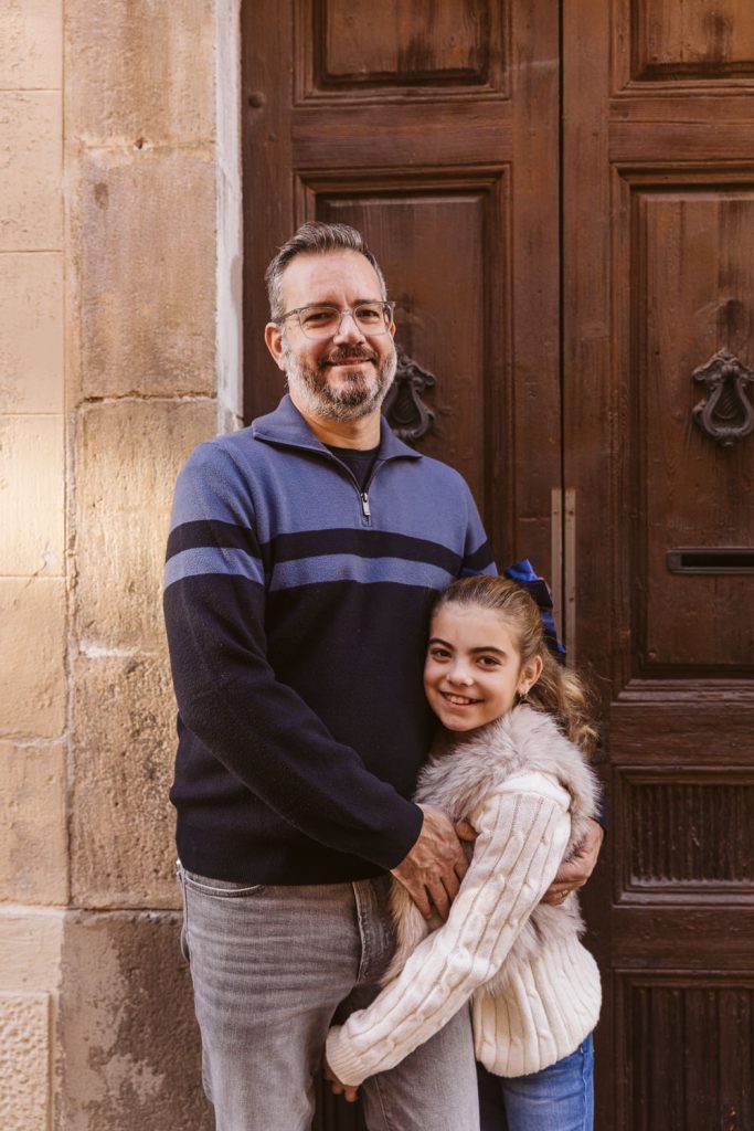 A man with glasses and a beard smiles while standing in front of a wooden door, hugging a young girl with long hair and a fur vest during a family photo session in Vila de Gràcia Barcelona.