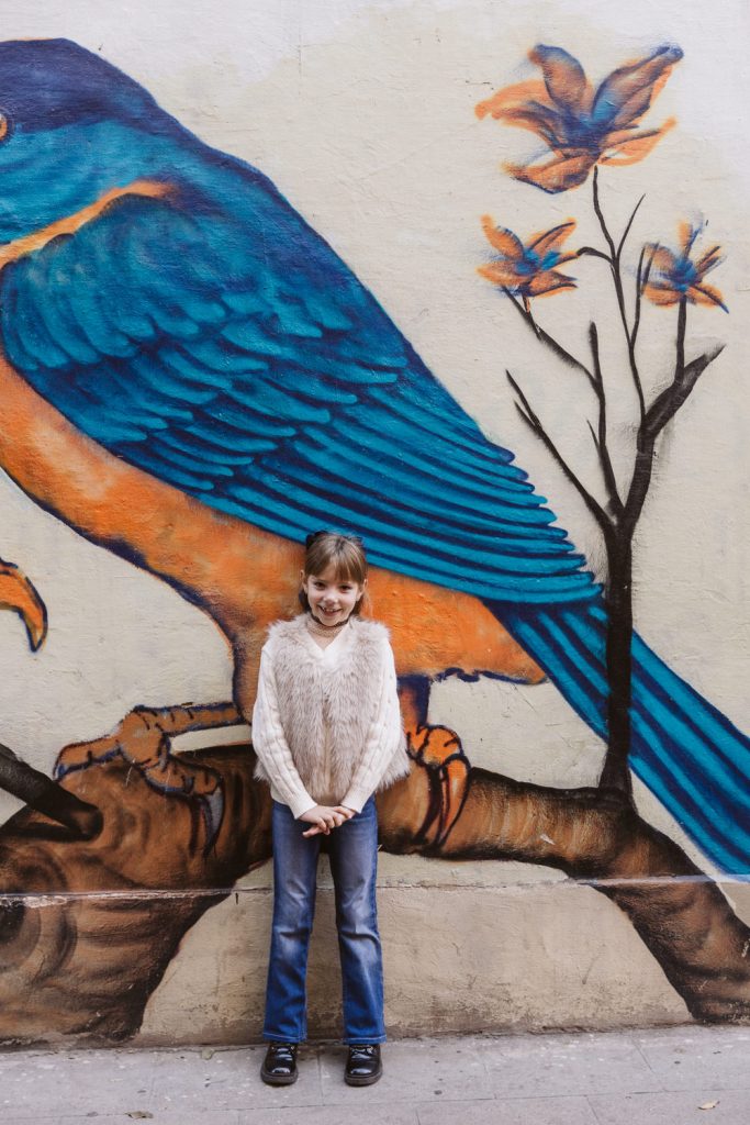 A young girl in jeans and a furry sweater stands smiling in front of a colorful mural featuring a large blue and orange bird and flowers—capturing a joyful moment from a family photo session in Vila de Gràcia, Barcelona.