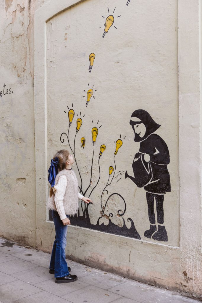 A girl with a blue ribbon in her hair looks up at a mural showing a figure watering plants with blooming light bulbs as flowers—a whimsical scene, perfect for a family photo session in Vila de Gràcia, Barcelona.