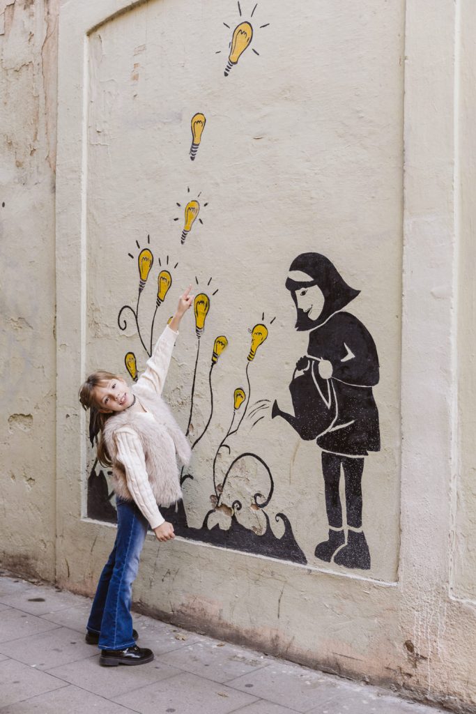 A young girl smiles and points at a mural on a wall during a family photo session in Vila de Gràcia, Barcelona. The mural shows a girl watering plants that are growing light bulbs, with more light bulbs floating upward.