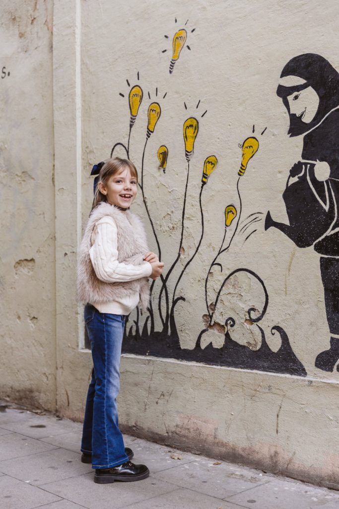 A smiling girl stands on a sidewalk in Vila de Gràcia, Barcelona, next to a mural of a figure watering flowers with yellow light bulbs blooming—capturing the magic of a family photo session in this vibrant neighborhood.