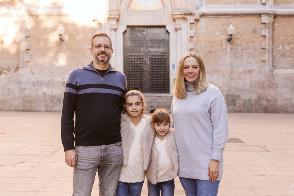 A family of four stands outdoors in front of an old stone building during a family photo session in Vila de Gràcia, Barcelona. The two adults and two children smile warmly at the camera, dressed in cozy sweaters and jeans.