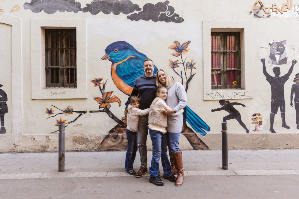 A family of four stands smiling in front of a mural featuring a large blue and orange bird, with flowers and painted figures—a cozy family photo session in Vila de Gràcia, Barcelona. The parents and two children wear sweaters and jeans.