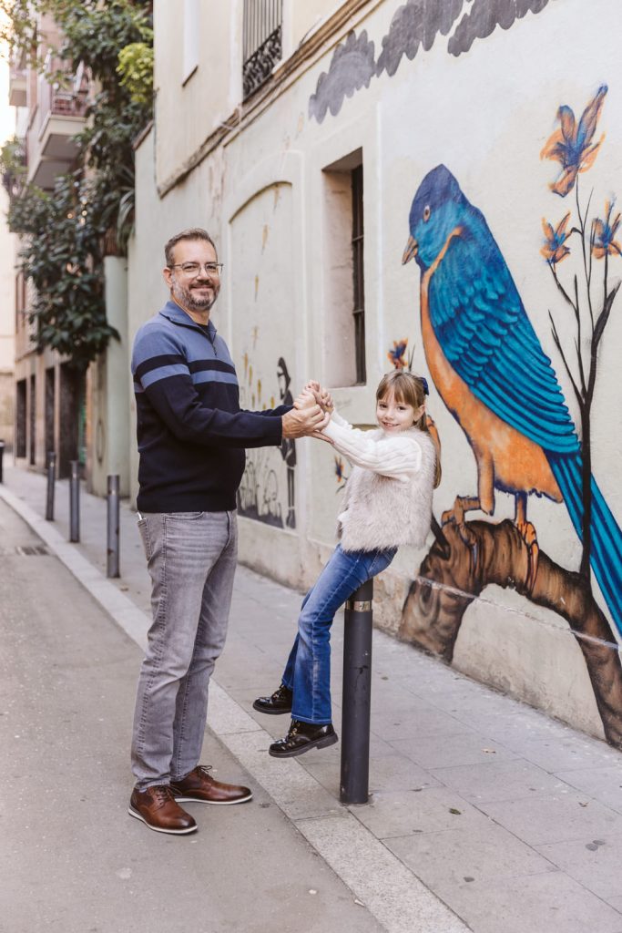 A man and a young girl hold hands and smile on a city sidewalk in front of a colorful mural featuring a large blue and orange bird—a perfect moment from a family photo session in Vila de Gràcia, Barcelona.