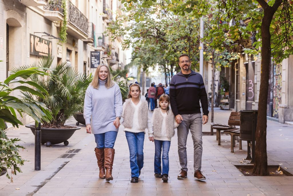 A family of four walks hand in hand down a tree-lined city street during their family photo session in Vila de Gràcia, Barcelona. The parents and two young girls are smiling, dressed in casual fall clothes as shops and greenery line the sidewalk.