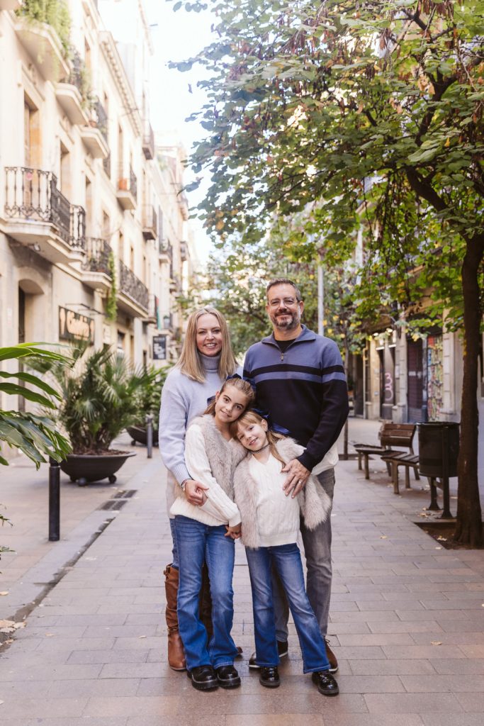 A smiling family of four—two adults and two young girls—stand close together on a tree-lined, sunlit street during a family photo session in Vila de Gràcia, Barcelona, with apartment buildings, plants, and benches in the background.