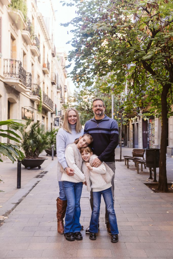 A smiling family of four, dressed in sweaters and jeans, hugs and poses for a family photo session in Vila de Gràcia Barcelona on a tree-lined street with charming buildings and benches in the background.