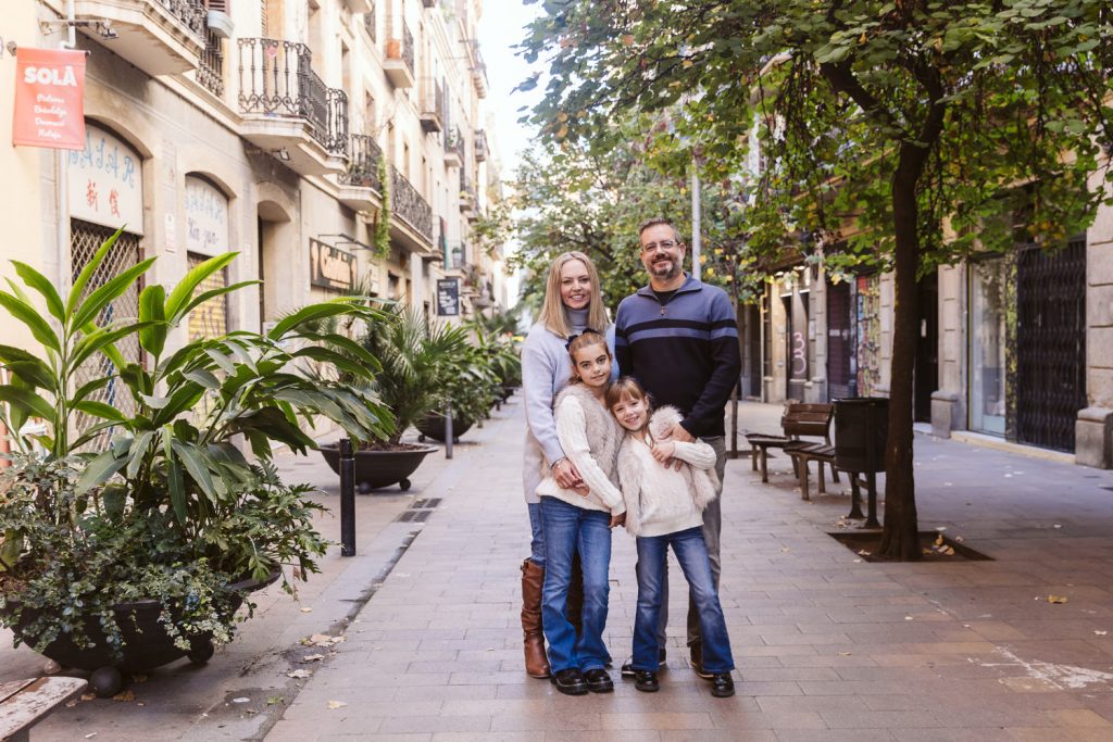 A family of four, including two young girls and their parents, stands smiling together on a tree-lined city street with plants and old buildings in the background during a family photo session in Vila de Gràcia, Barcelona.