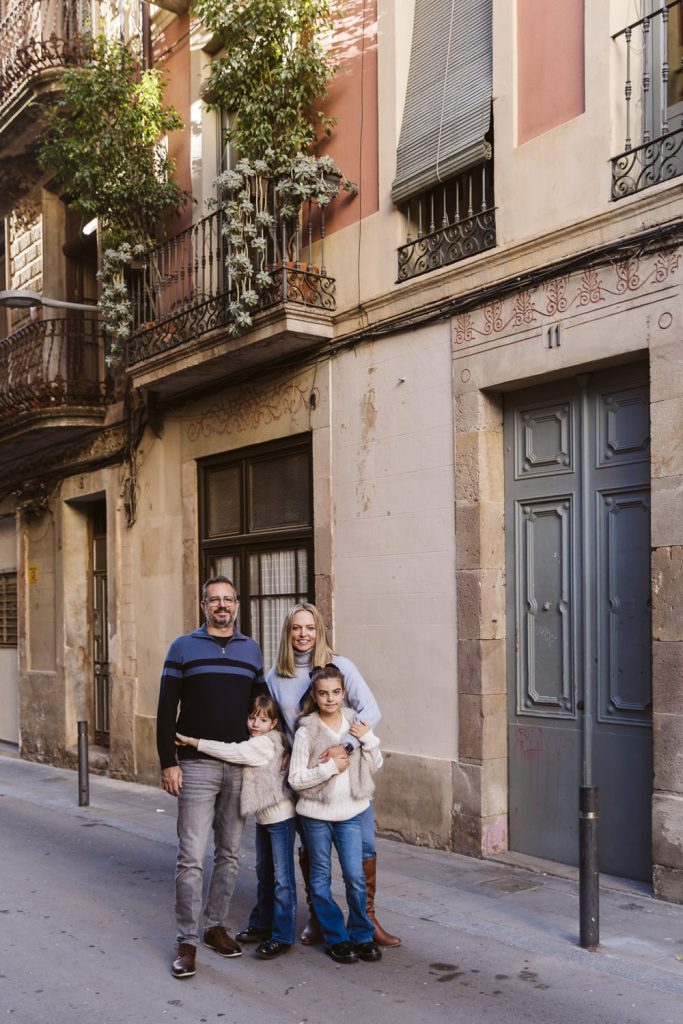 A family of four stands close together, smiling in front of an old building with balconies and green plants—a perfect moment from a family photo session in Vila de Gràcia, Barcelona. The adults and two children wear casual sweaters and jeans.