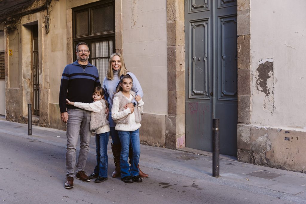 A family of four stands together on a narrow street by an old building during a family photo session in Vila de Gràcia, Barcelona. The parents smile with their two children in front, all wearing sweaters and jeans, posing happily for the photo.