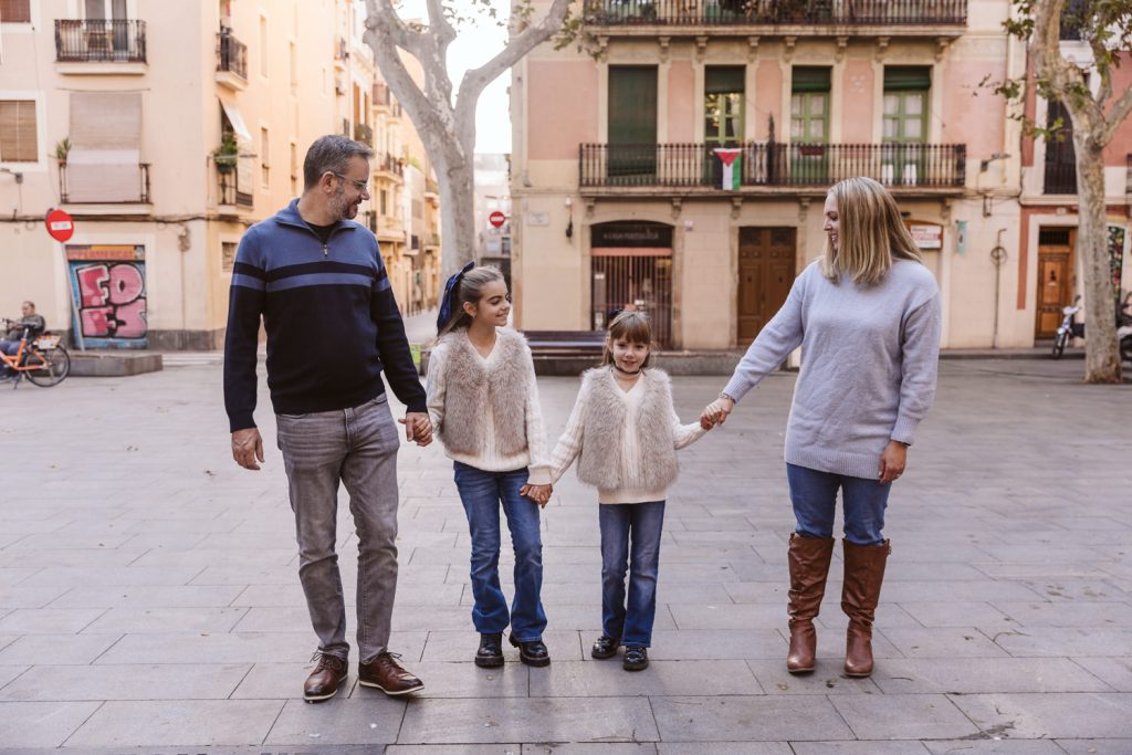 A family of four stands holding hands in a city square during a family photo session in Vila de Gràcia, Barcelona. Two young girls are in the middle with their parents on either side, all casually dressed and enjoying time together outdoors.