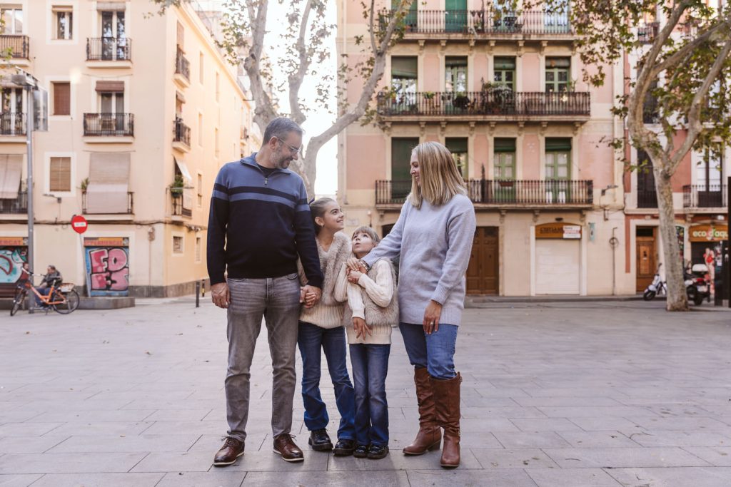 A family of four stands together in an urban plaza during a family photo session in Vila de Gràcia, Barcelona, holding hands and smiling at each other in front of apartment buildings with trees nearby.
