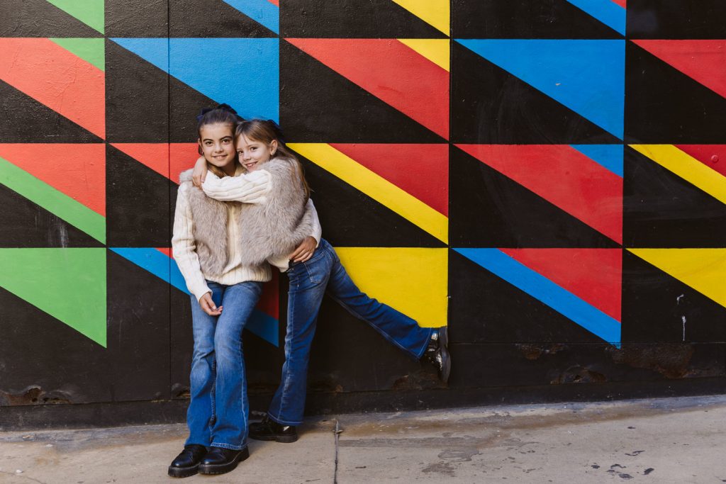 Two young girls in matching outfits—furry vests, sweaters, and flared jeans—smiling and posing during a family photo session in Vila de Gràcia Barcelona, in front of a colorful mural with red, yellow, and blue triangles.