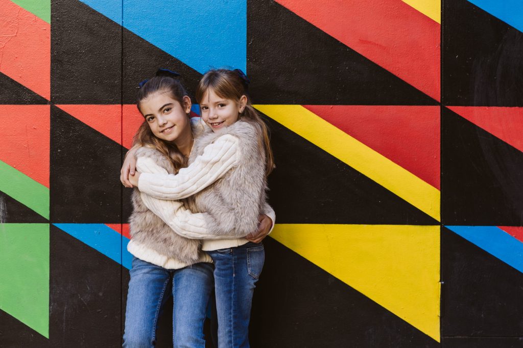 Two young girls smiling and hugging each other during a family photo session in Vila de Gràcia, Barcelona, standing in front of a colorful geometric mural with red, blue, yellow, and black triangles. Both wear light sweaters and jeans.