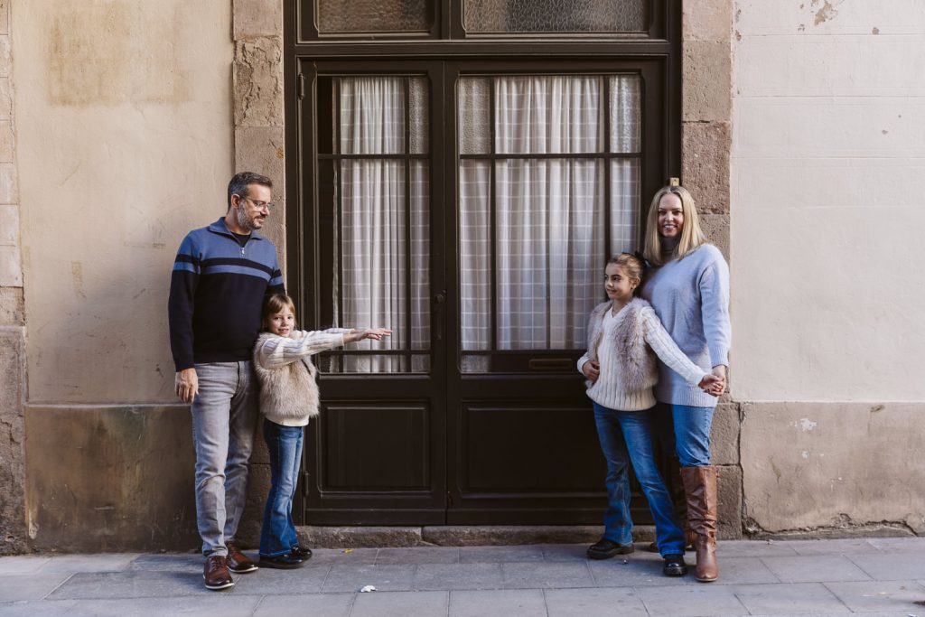A family photo session in Vila de Gràcia, Barcelona: A family of four poses by a large window with curtains. The father and son stand on the left, while the mother and daughter hold hands on the right. The son playfully stretches his arms toward his sister.