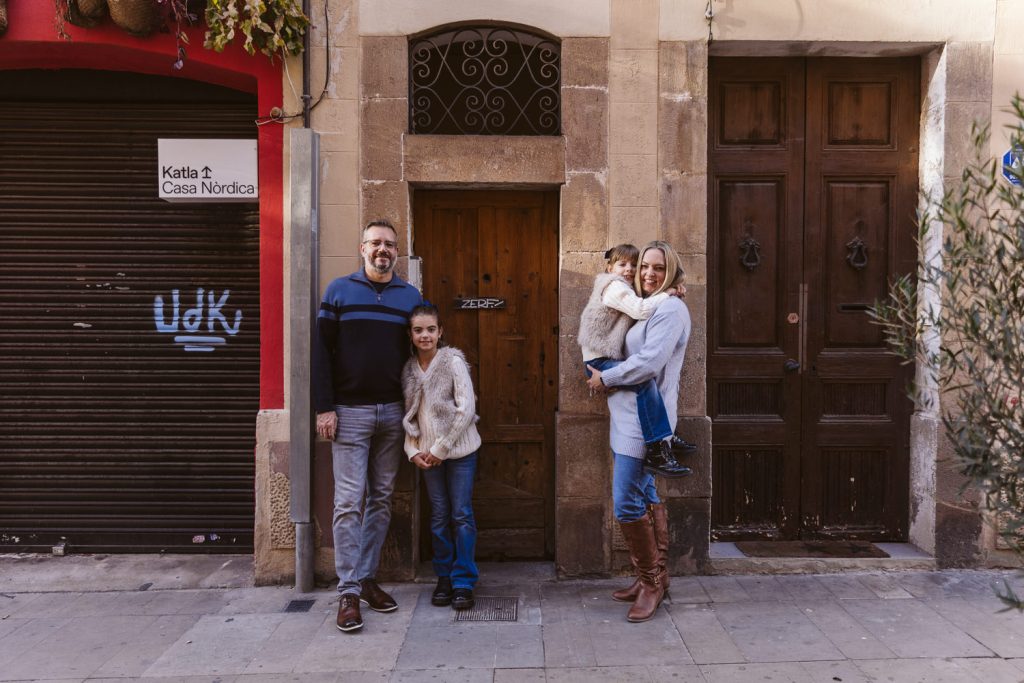 A family photo session in Vila de Gràcia Barcelona: a family of four poses on a city sidewalk in front of wooden doors, dressed in casual, warm clothing. A sign reads Katta Casa Nordica.