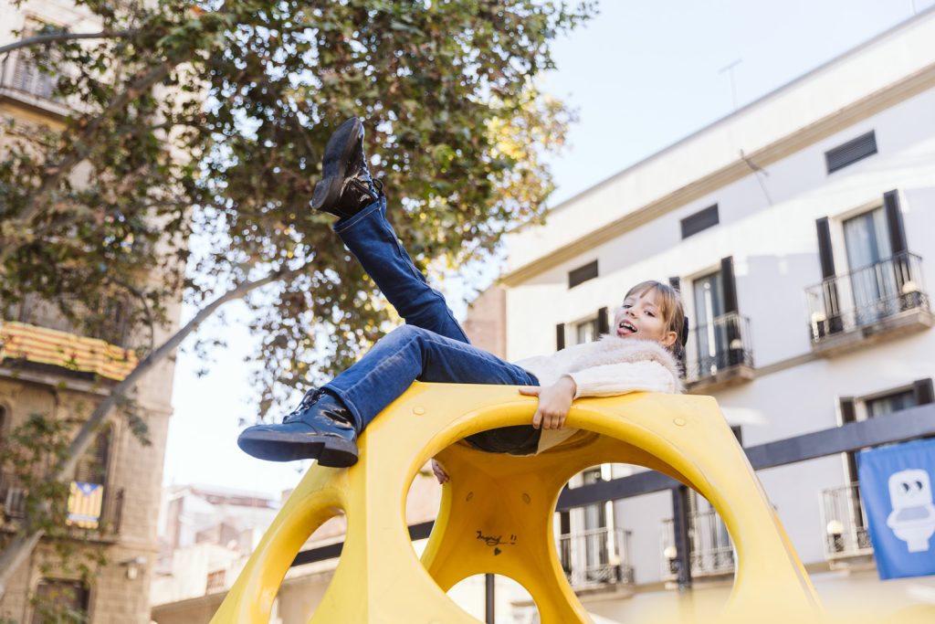 A young child in jeans and a white sweater smiles while playfully reclining on top of a yellow playground structure during a family photo session in Vila de Gràcia, Barcelona. Buildings and leafy trees are visible in the background.