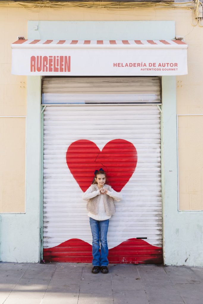 A girl stands in front of a shop with a heart painted on its metal shutter, making a heart shape with her hands—capturing the warmth of a family photo session in Vila de Gràcia, Barcelona. The awning reads “Aurelien Heladería de Autor.”.