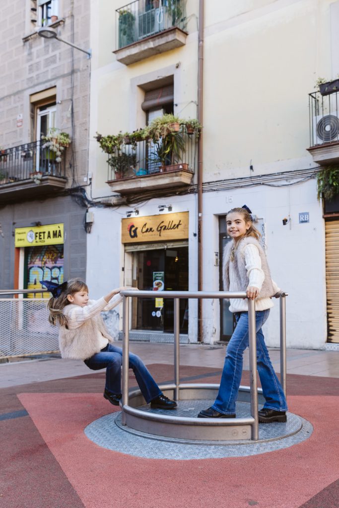 Two young girls wearing light sweaters and jeans play on a metal merry-go-round in an urban plaza, perfect for a family photo session in Vila de Gràcia Barcelona, with storefronts and apartments visible in the background.