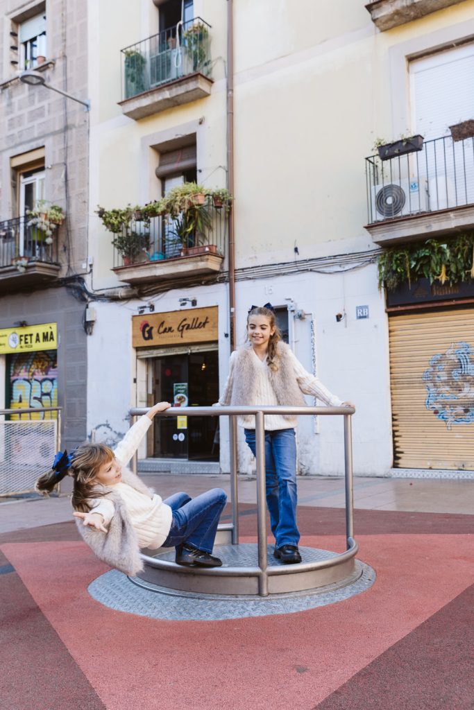 Two girls play on a roundabout in an urban playground during a family photo session in Vila de Gràcia Barcelona. One girl stands and spins the equipment while the other sits, leaning back and smiling. Apartment buildings with balconies are visible behind them.