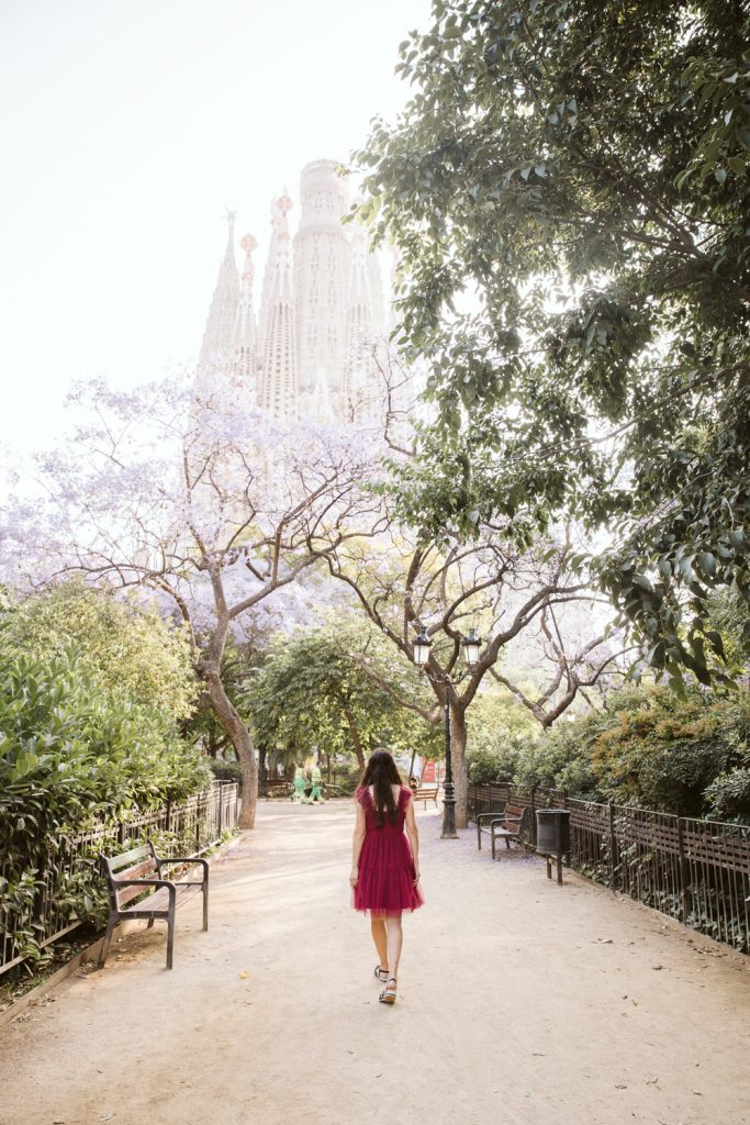 A woman in a pink dress walks along a tree-lined path toward the Sagrada Familia in Barcelona, with purple blossoms and park benches on either side.
