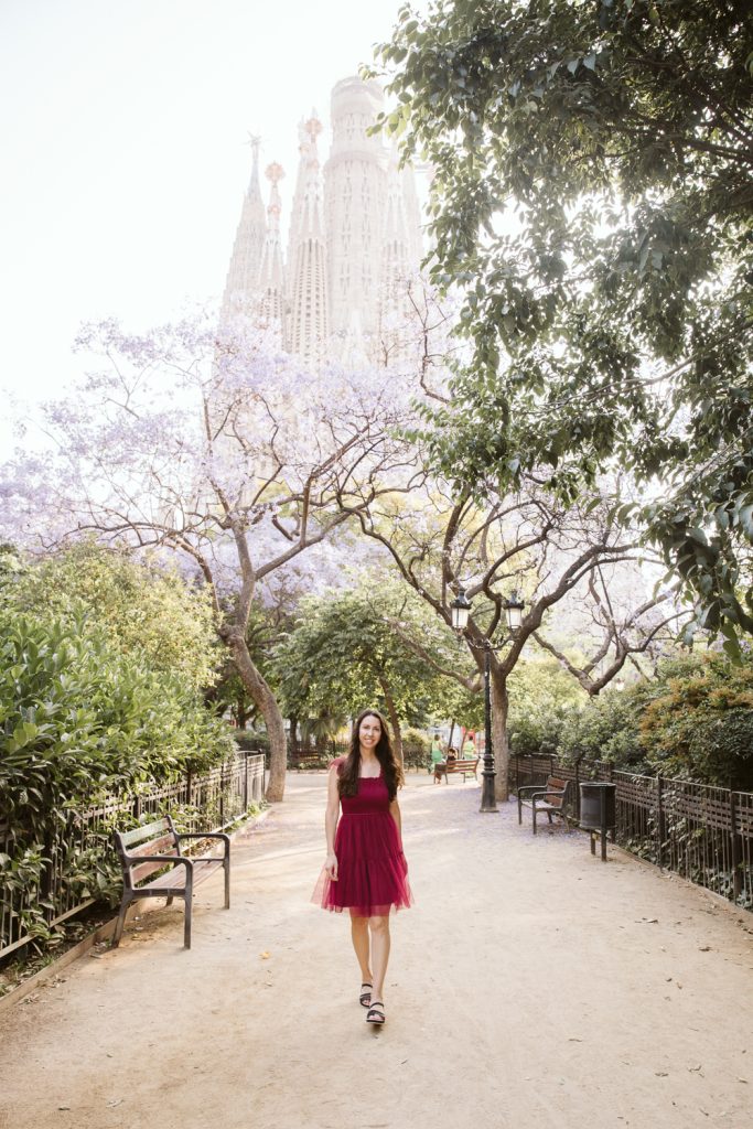 A woman in a red dress walks along a sunlit path lined with benches and trees, with the Sagrada Família basilica rising in the background in Barcelona.